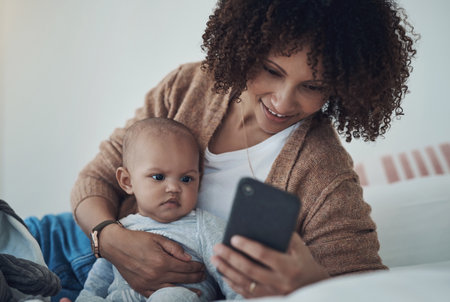 An awesome new app for a new mom. a young woman using a smartphone while relaxing on the bed with her adorable baby girl at home.の写真素材