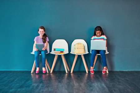 Supporting schoolwork with wireless technology. Studio shot of kids sitting on chairs and using wireless technology with clouds above them against a blue background.の写真素材