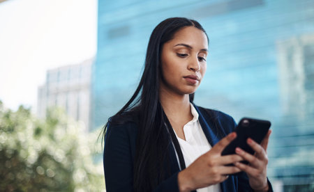 Available on the move thanks to mobile technology. a young businesswoman using a smartphone against a city background.の写真素材