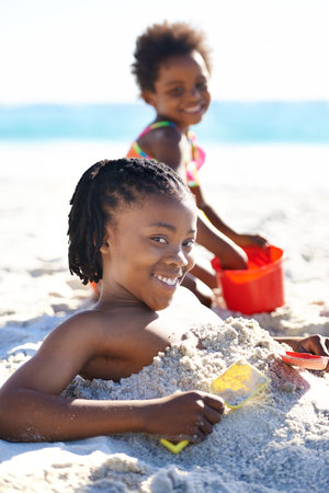 Hes going to have to dig himself out again. A young boy burying himself in beach sand as his sister watches.の写真素材