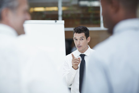 Caution in the work place. A handsome businessman stands in front of a flipchart pointing in the direction of colleagues in the foreground.の写真素材