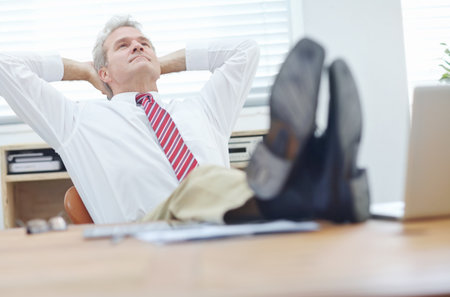 Thinking about tomorrows challenges. Mature businessman with his feet up on a desk leaning back in an office chair with his hands behind his head.の写真素材