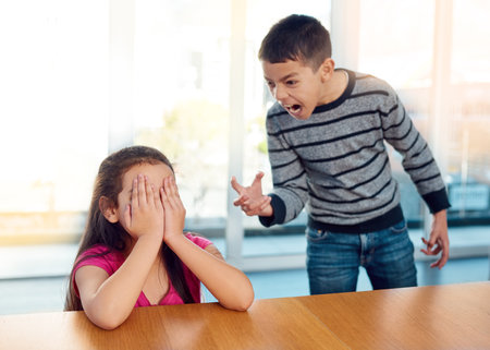 They drive each other crazy. a little girl covering her face while her brother angrily screams at her at home.の写真素材