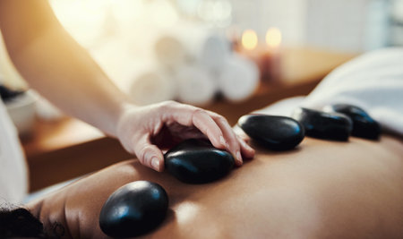 Woman, hands and rocks in relax for back massage, skincare or beauty relaxation on bed at spa. Hand of masseuse applying hot rock or stones on female for physical therapy or treatment at a resortの写真素材