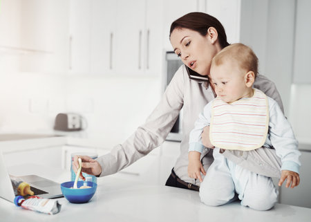 Family, baby and mother with multitasking, home and phone call with stress, feeding toddler and burnout. Mama, female parent and infant in the kitchen, smartphone and communication with frustrationの写真素材