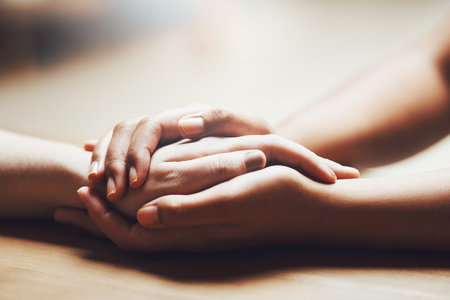 Prayer, empathy and support with people holding hands in comfort, care or understanding on a wooden table of a home. Love, faith or depression with friends closeup for help, hope or peace for healingの写真素材