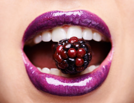Closeup of woman lips with purple lipstick, makeup and beauty with shine and vibrant aesthetic, creativity and glow. Fruit between female model teeth, cosmetic product for mouth and cosmetologyの写真素材