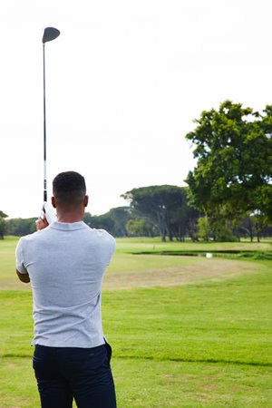 All about the follow through. Rear view shot of a young man who has just hit a golf ball.の写真素材