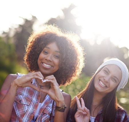 Peace, love and happiness. A young woman make a heart shape with her hands while standing beside her friend outside.の写真素材