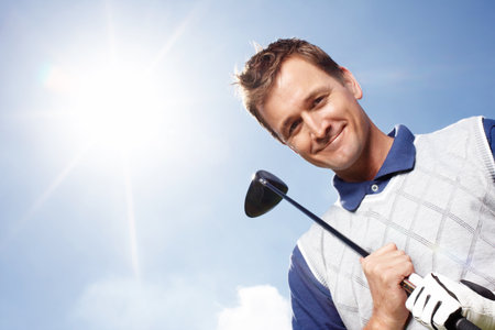 Want to play some golf. Smiling mature man holding his golf club while against a blue sky - low angle.の写真素材
