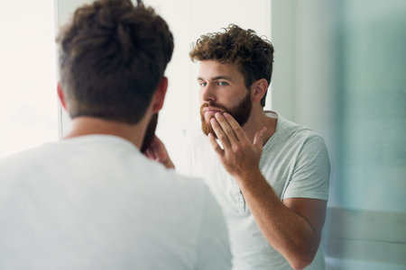 Skincare, beauty and beard with a man in the bathroom of his home in the morning for fresh hygiene. Mirror, reflection and self care with a handsome young male person checking his face or skinの写真素材