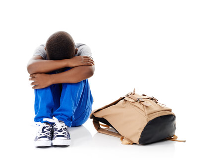 I dont want to. Studio shot of a little boy with his head buried in his knees sitting next to his schoolbag against a white background.の写真素材