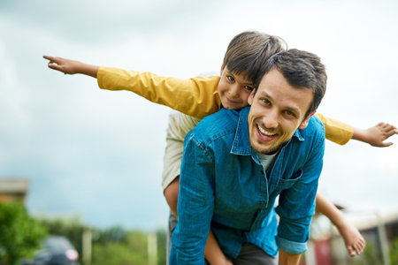 Portrait, children and a son on back of his dad outdoor in the garden to fly like an airplane while bonding together. Family, kids and a father carrying his boy child while playing a game in the yardの写真素材