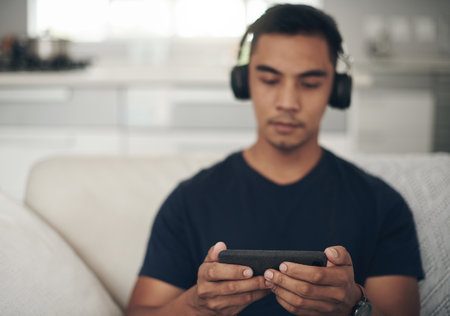 Glued to his screen for the weekend. a young man wearing headphones and using a cellphone while relaxing at home.の写真素材