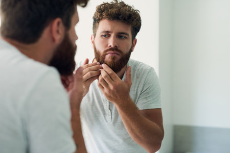 Face, beauty and beard with a man in the bathroom of his home in the morning for fresh hygiene. Mirror, reflection and care with a handsome young male person checking his skin for facial treatmentの写真素材