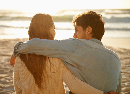 Waiting for the sun to set...Rear-view shot of a young couple sitting on the beach and sharing a romantic moment.の写真素材