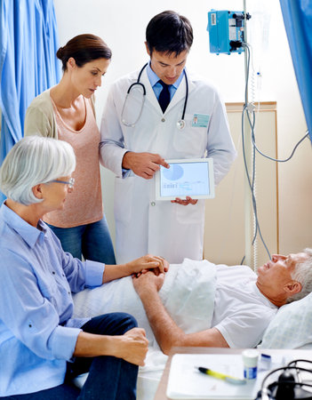 This could get rather graphic. a sick man in a hospital bed being shown a graph on a tablet while his wife holds his hand and his daughter looks on.の写真素材