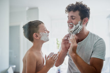 Kid, father and learning to shave, funny or bonding together in home bathroom. Laughing, dad and teaching child with shaving cream on face beard, playing or cleaning, hygiene or enjoying hair removalの写真素材