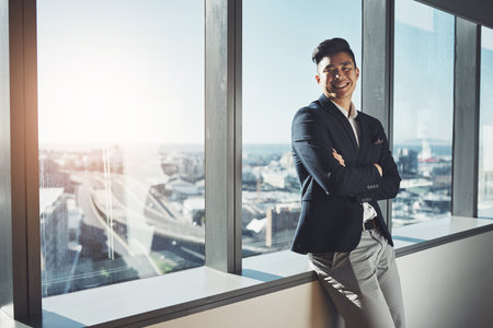 Hes a leading executive in the business empire. Portrait of a young businessman standing at a window in an office.の写真素材