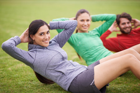 Working out together makes us push ourselves more. Portrait of three friends doing sit-ups together on a sports field.の写真素材