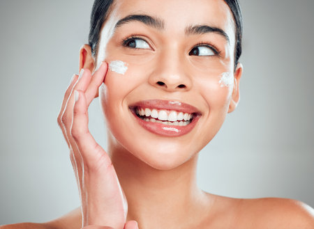 A beautiful smiling happy mixed race woman applying cream to her face. Hispanic model with glowing skin against a grey copyspace backgroundの写真素材