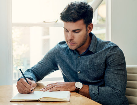 Note-taking is essential for success. a handsome young businessman sitting and writing in a notebook while in the office alone.の写真素材