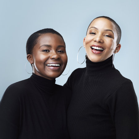 Yes we are wearing the same jersey. Portrait of two beautiful young women holding each other while standing against a grey background.の写真素材