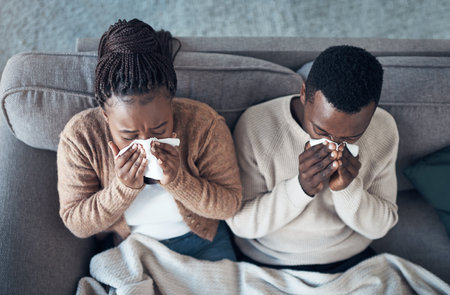 At least were sick together. High angle shot of a young couple sitting in their living room together and feeling sick while blowing their nose.の写真素材