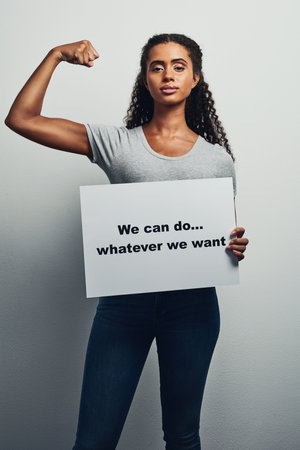 Who says you not powerful. Studio shot of an attractive young woman holding a placard that reads We can do whatever we want against a grey background.の写真素材