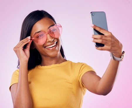 Sunglasses, selfie and happy Indian woman on pink background with cosmetics, confidence and beauty. Fashion, summer style and girl in studio take picture for social media, post and influencer blogの写真素材