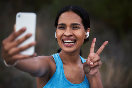 Selfie, peace and fitness with a sports woman outdoor, taking a picture during her cardio or endurance workout. Exercise, running and smile with a happy young female athlete posing for a photographの写真素材