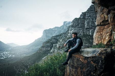 The man on top of the mountain didnt fall there. a young man looking at the view while sitting on a mountain cliff.の写真素材