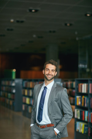 Educate yourself at any chance you get. Cropped portrait of a handsome young businessman standing with his hands in his pockets in an empty library.の写真素材