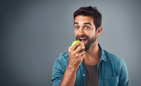 Real men eat real food. Studio shot of a handsome young man eating an apple against a grey...
