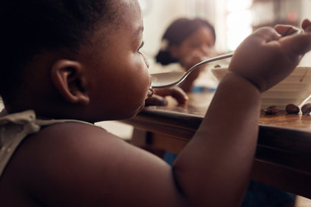 Black child, cereal spoon and eating baby in a home kitchen with food and bowl at breakfast. African girl, nutrition and youth in a house with hungry kid relax with healthy snack and kids at morningの写真素材