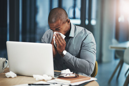 If only I could fend off this flu. a young businessman blowing his nose while working in an office.の写真素材
