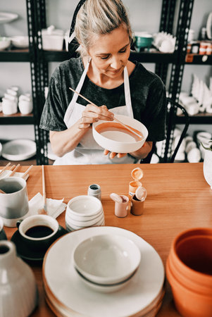 Creativity is intelligence having fun. an attractive mature woman sitting alone and painting a pottery bowl in her workshop.の写真素材