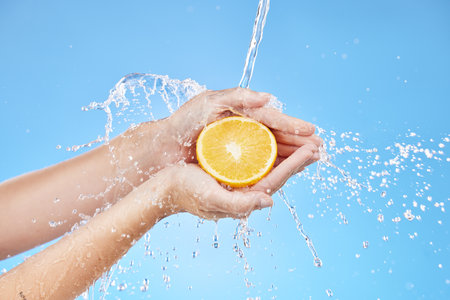 Water splash, zoom or orange in hands for skincare wellness, nutrition or health in blue background studio. Water, shower or cleaning fruit in hand for natural, organic or fresh food vitamin C splashの写真素材