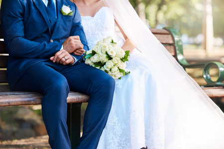 Closeup of a bride and groom sitting on a park bench together. Man wearing suit and woman wearing wedding dress and holding bouquet while sitting outdoorsの写真素材