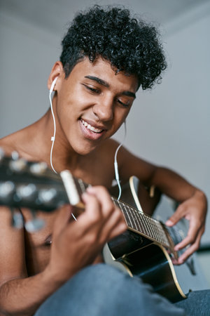 The best time to build your skills is now. a young man using earbuds while playing the guitar at home.の写真素材