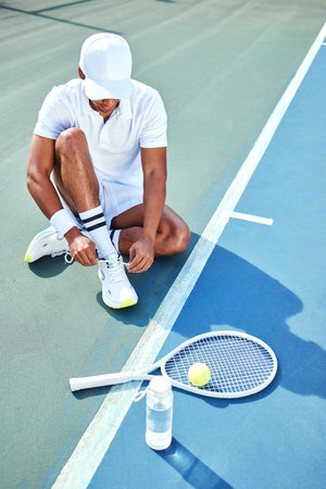 Getting ready for a long training session. Full length shot of an unrecognisable man sitting alone and tying his shoelaces before tennis practice.の写真素材