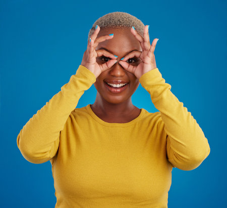 Black woman, binocular hands and eyes in studio portrait for comic funny face with smile, fashion and happy. Student girl, model and ok hand gesture with happiness, youth and pride by blue backgroundの写真素材