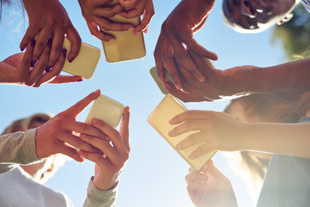 Hands, low angle and phone by friends together for social media, networking and sharing app on blue sky background. Below, smartphone and gen z people with internet, addiction or online communicationの写真素材