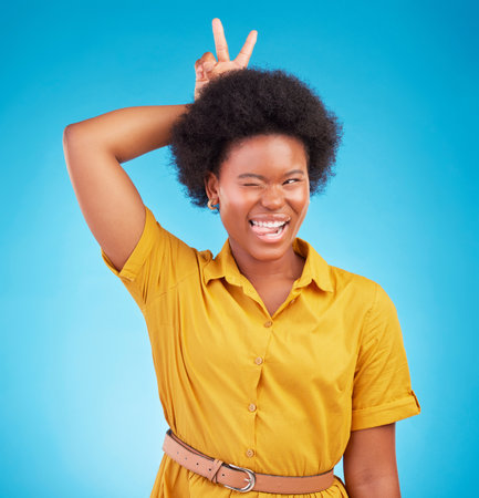 Silly, peace sign and woman in a studio feeling fun, playful and funny with a blue background. V hand gesture, bunny ears and tongue out for comedy with a young female model smile with happinessの写真素材