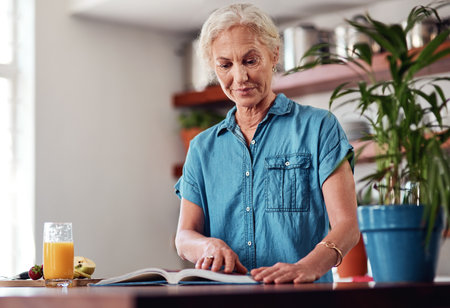 This looks like a goodie...an attractive senior woman reading from a recipe book while preparing breakfast in the kitchen.の写真素材