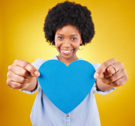 Paper, heart and portrait of black woman in studio for romance, positive and emotion. Happiness, love and giving with female isolated on yellow background for date, feelings and affectionateの写真素材