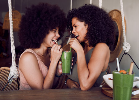 Friends, woman and drinking smoothie together in cafe, restaurant or date with comic laugh at table. Black women, share or smile with healthy juice, drink or health cocktail for love, bonding or careの写真素材