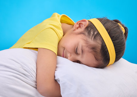 Calm young girl, pillow and sleep in a studio feeling tired, fatigue and ready for dreaming. Isolated, blue background and happy little child with pillows and closed eyes for sleeping, rest and napの写真素材