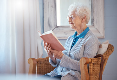 Book, reading and senior woman in home retirement with glasses and calm mindset, knowledge and learning on sofa chair. Elderly or old woman with literature, language and love story in her living roomの写真素材