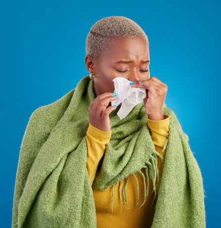 Sick black woman, tissue and blowing nose in studio, blue background or hayfever allergies, cold or winter virus. Female model, sneeze and influenza allergy of health problem, covid risk or sinusitisの写真素材